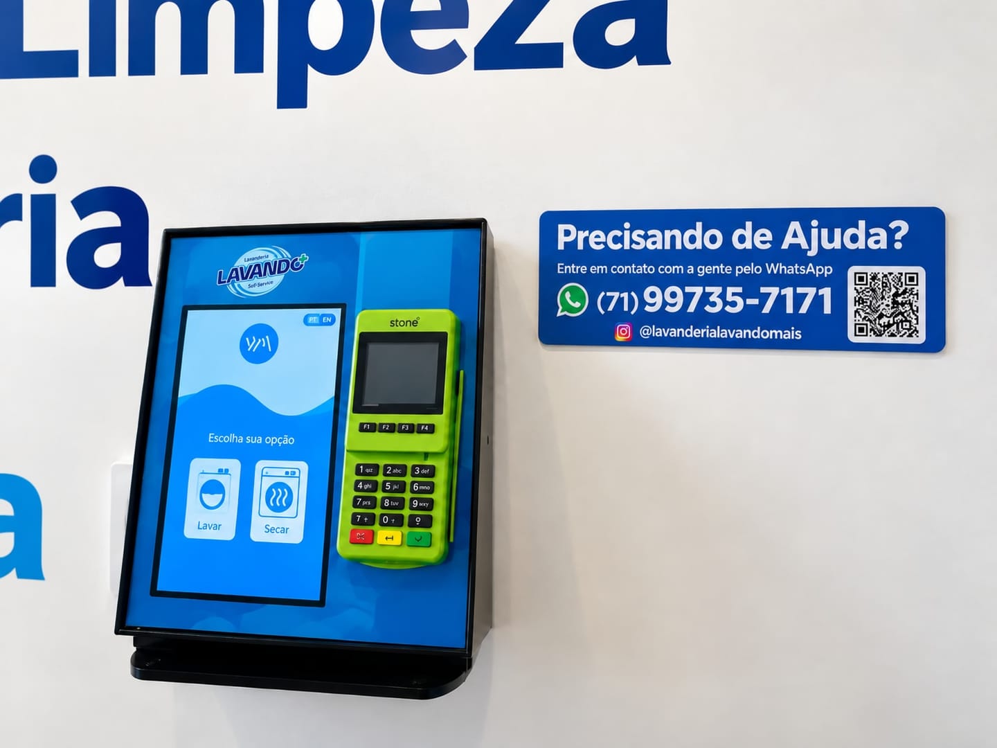 O totem de pagamento na parede, separado das máquinas: a tela Escolha sua opção com os botões Lavar e Secar, a maquininha de cartão ao lado e a placa Precisando de ajuda com o WhatsApp da loja